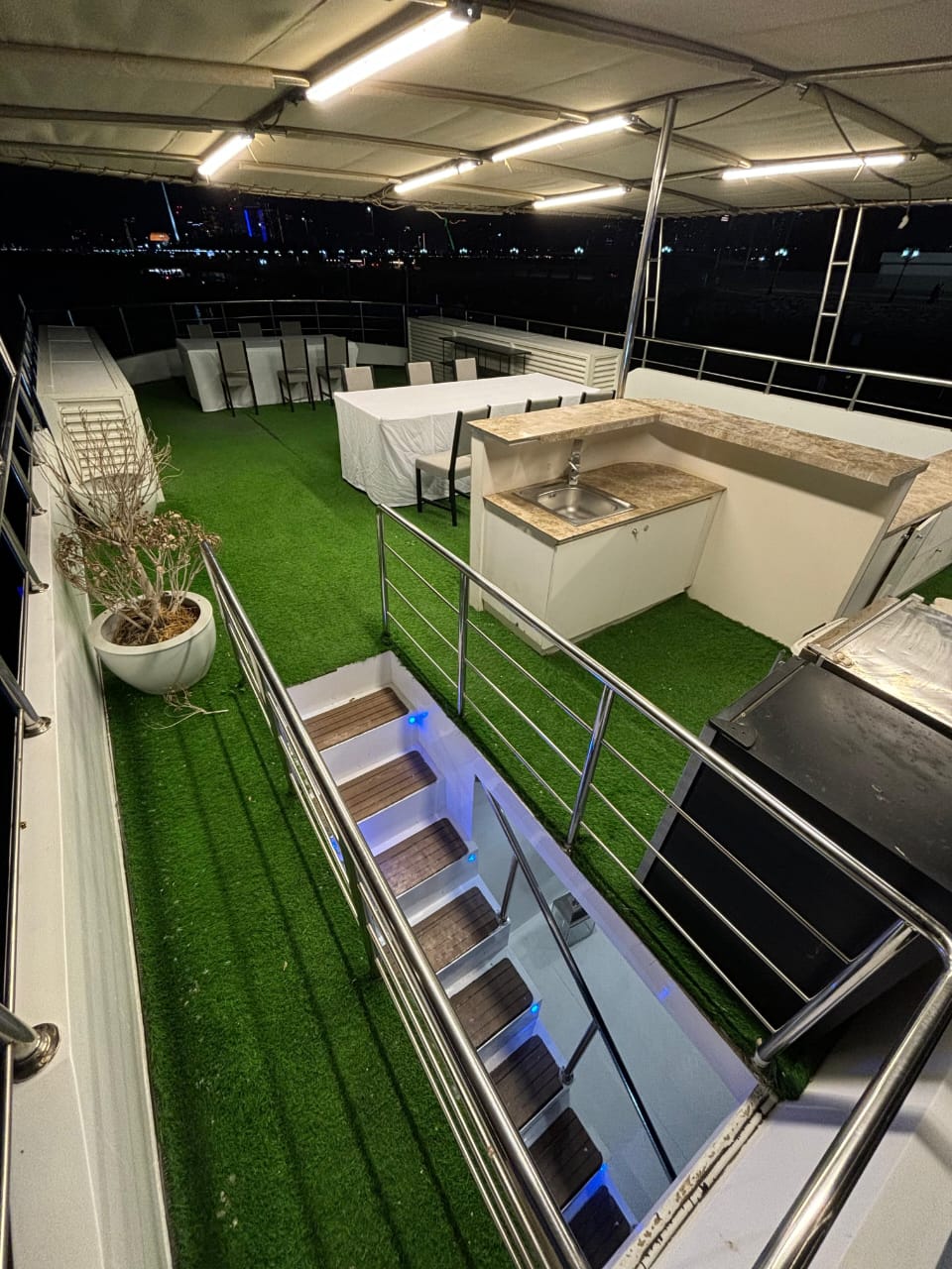 Upper deck at night with artificial grass, dining tables and a bar counter under a lit canopy