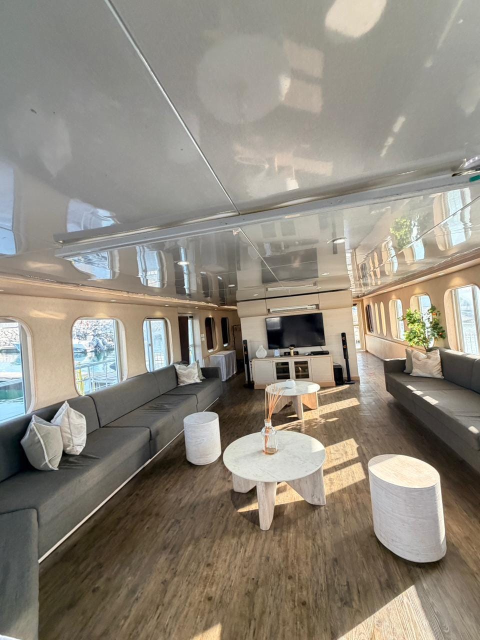 Long indoor lounge with grey sofas, marble coffee tables and a TV with speakers