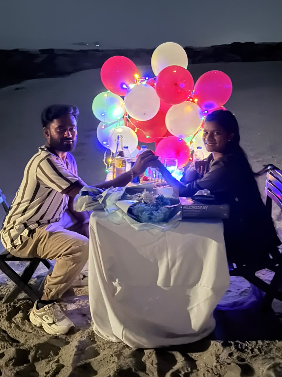 Couple holding hands at a candlelit birthday table set up on an Abu Dhabi beach with red and white balloons