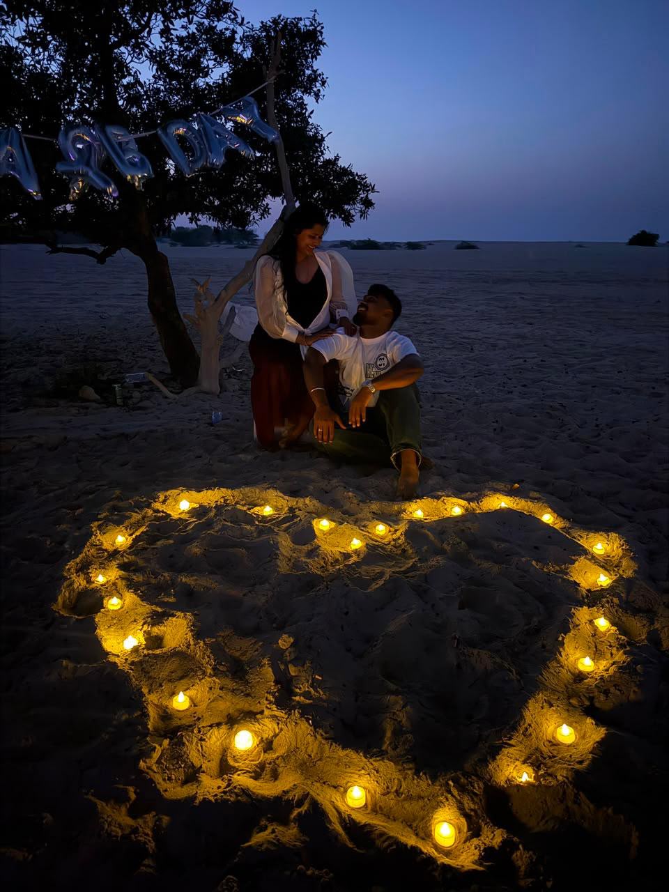 Couple sitting together beside a heart-shaped candle display on the sand at dusk