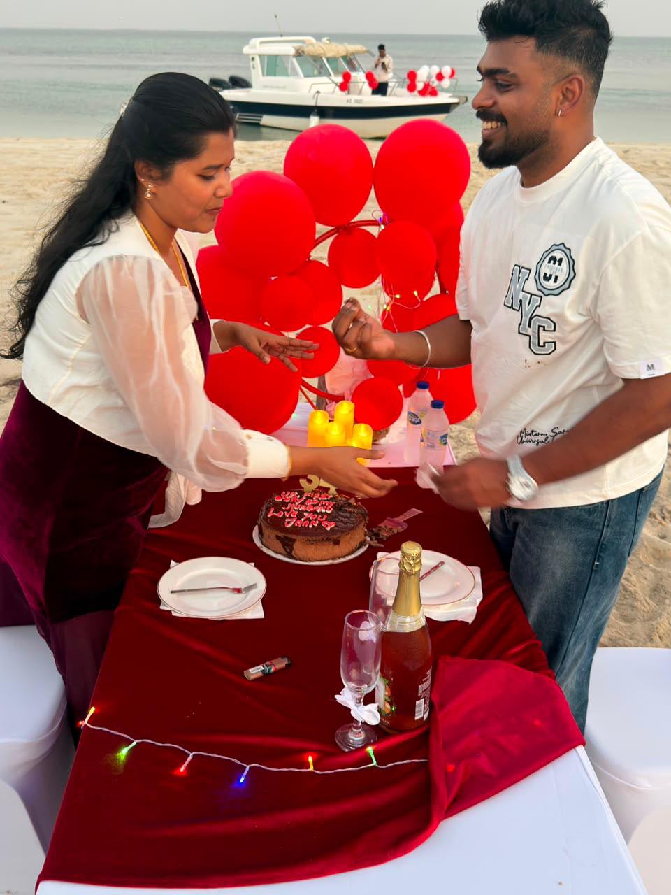 Couple cutting a birthday cake on the beach with red balloon decorations and the boat in the background
