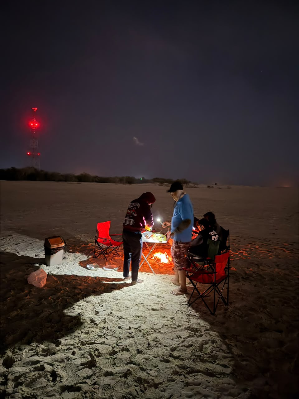 Group preparing a beach bonfire meal at night with a red-lit communications tower in the distance
