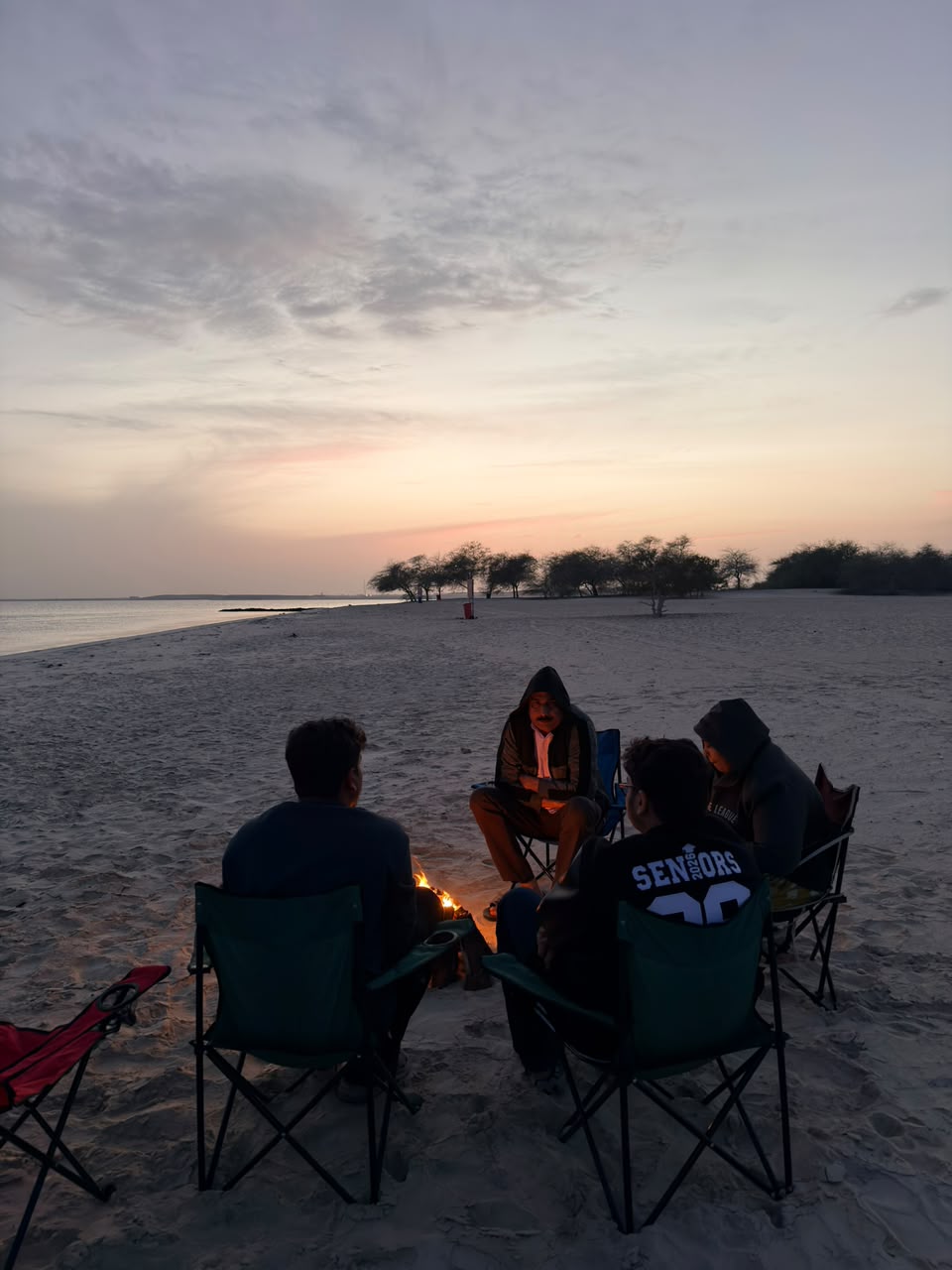 Group sitting around a bonfire on the beach at sunset with the sea and trees in the background
